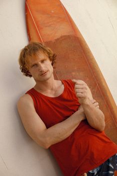 Casual portrait of a young man leaning against a surfboard wearing a red tank top