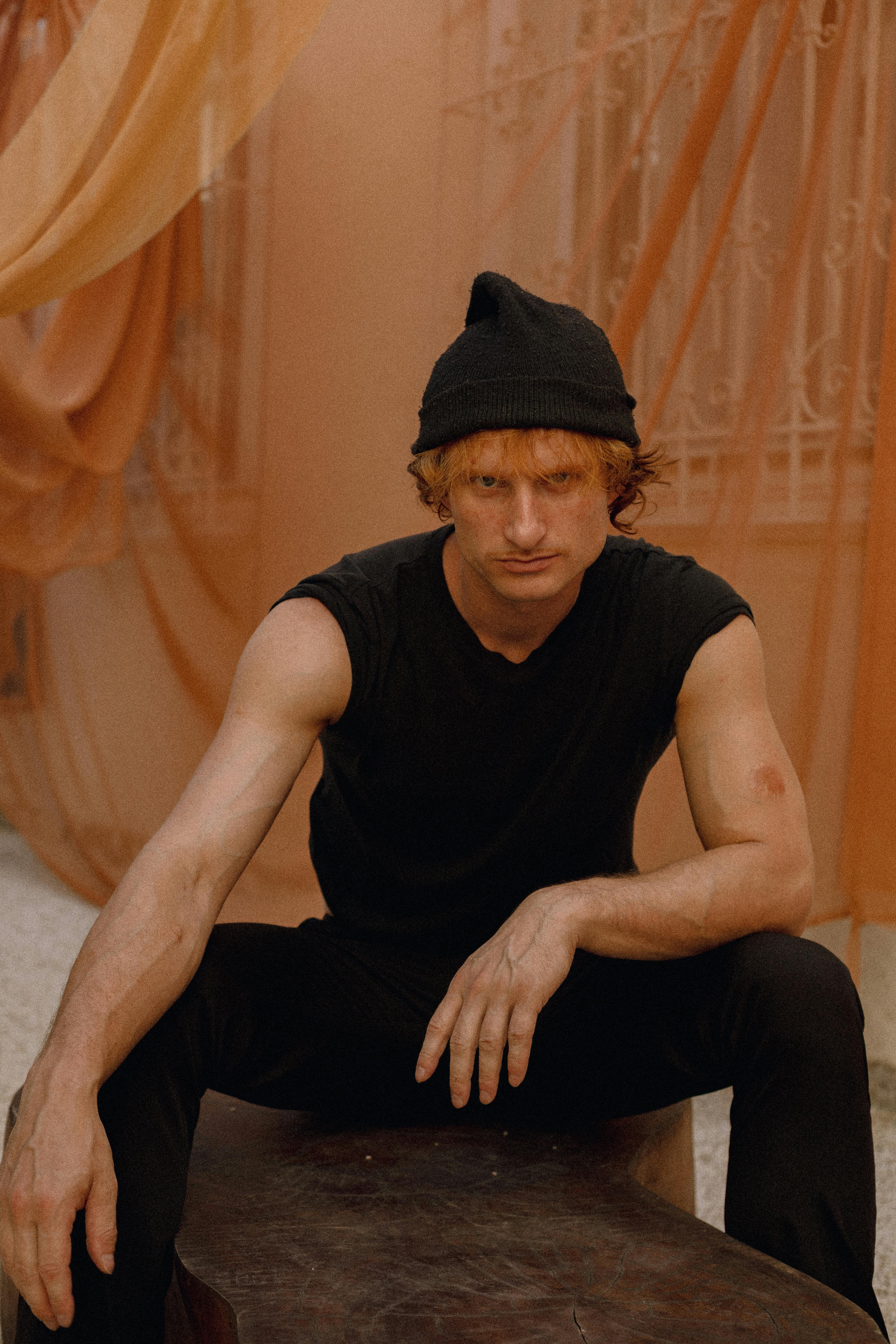 Man wearing black beanie and sleeveless shirt, sitting indoors with orange drapery background.