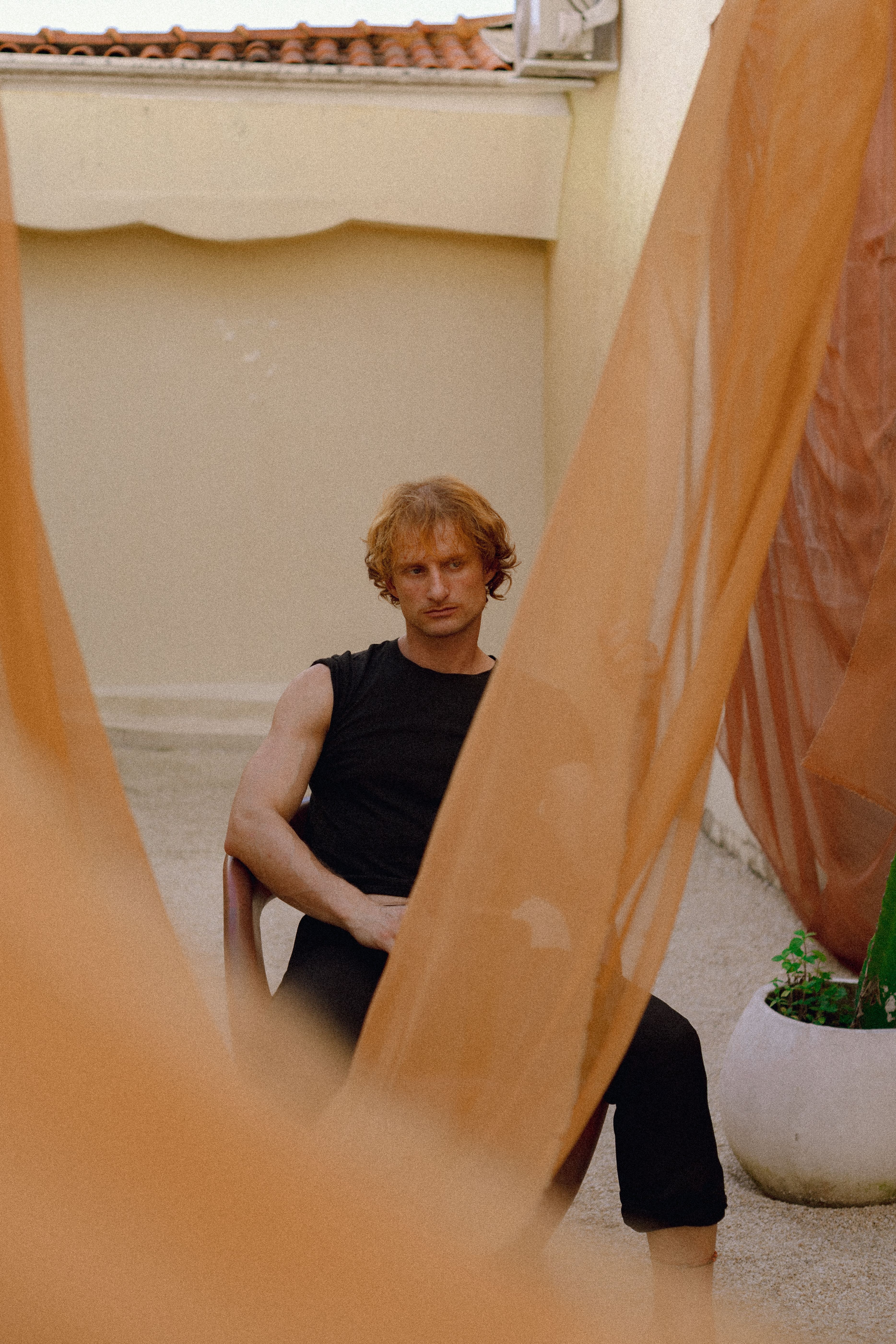 A man in black sits thoughtfully behind flowing sheer orange drapes in a courtyard setting.