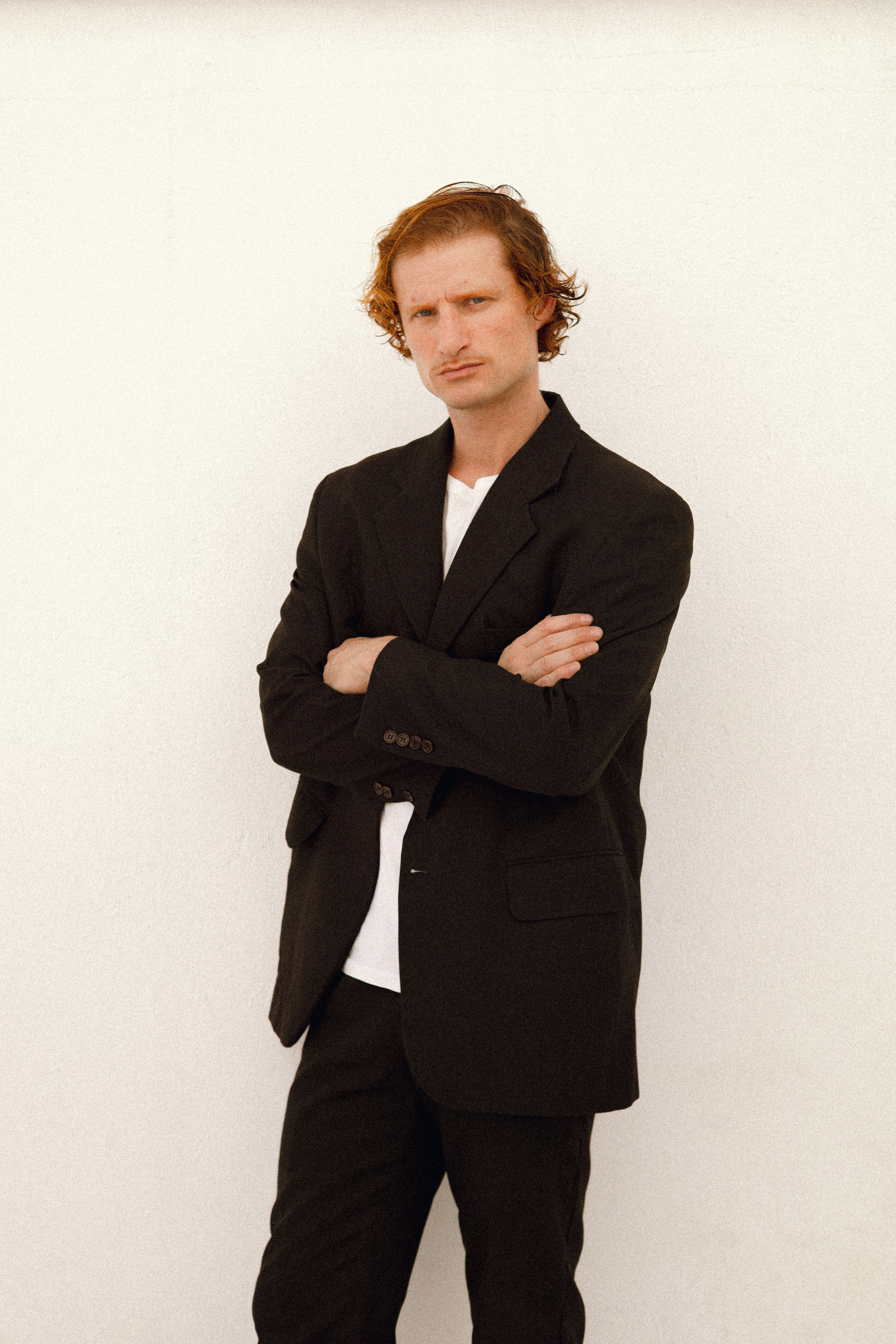 A man in a black suit stands confidently with arms crossed against a white backdrop.