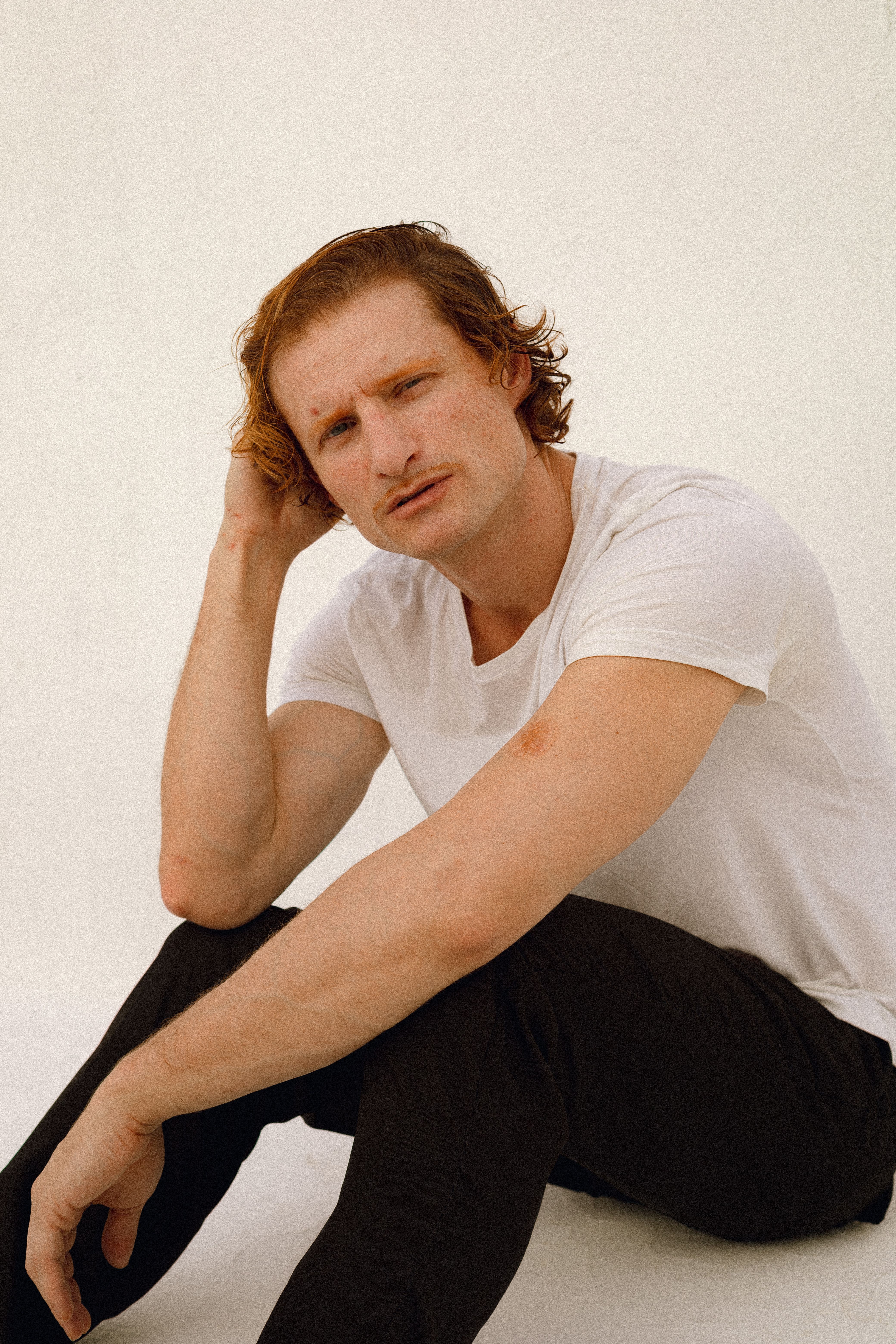 A casual portrait of a man in a white t-shirt, sitting thoughtfully indoors against a plain background.