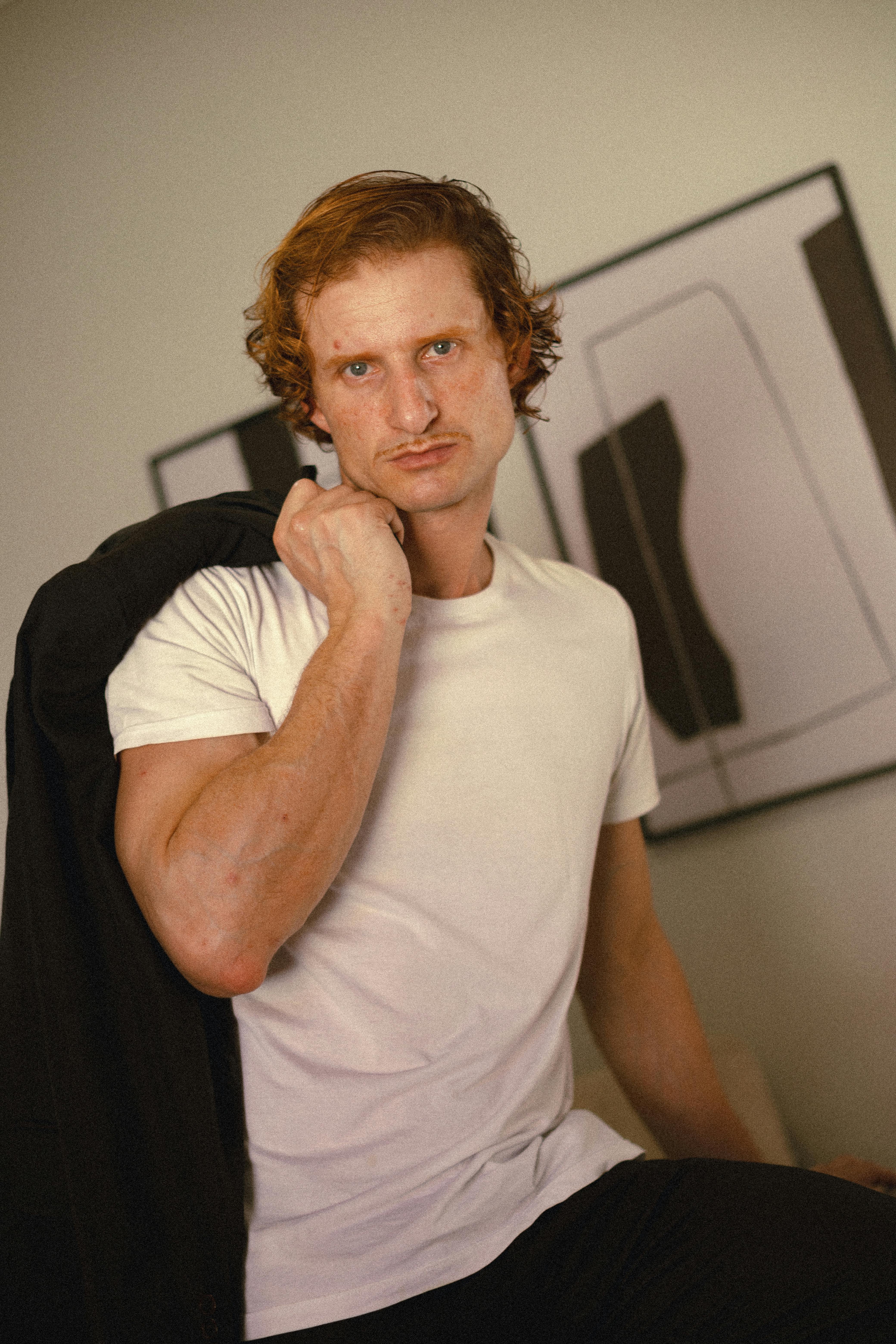 Man in a white t-shirt holding a jacket indoors with abstract art background.