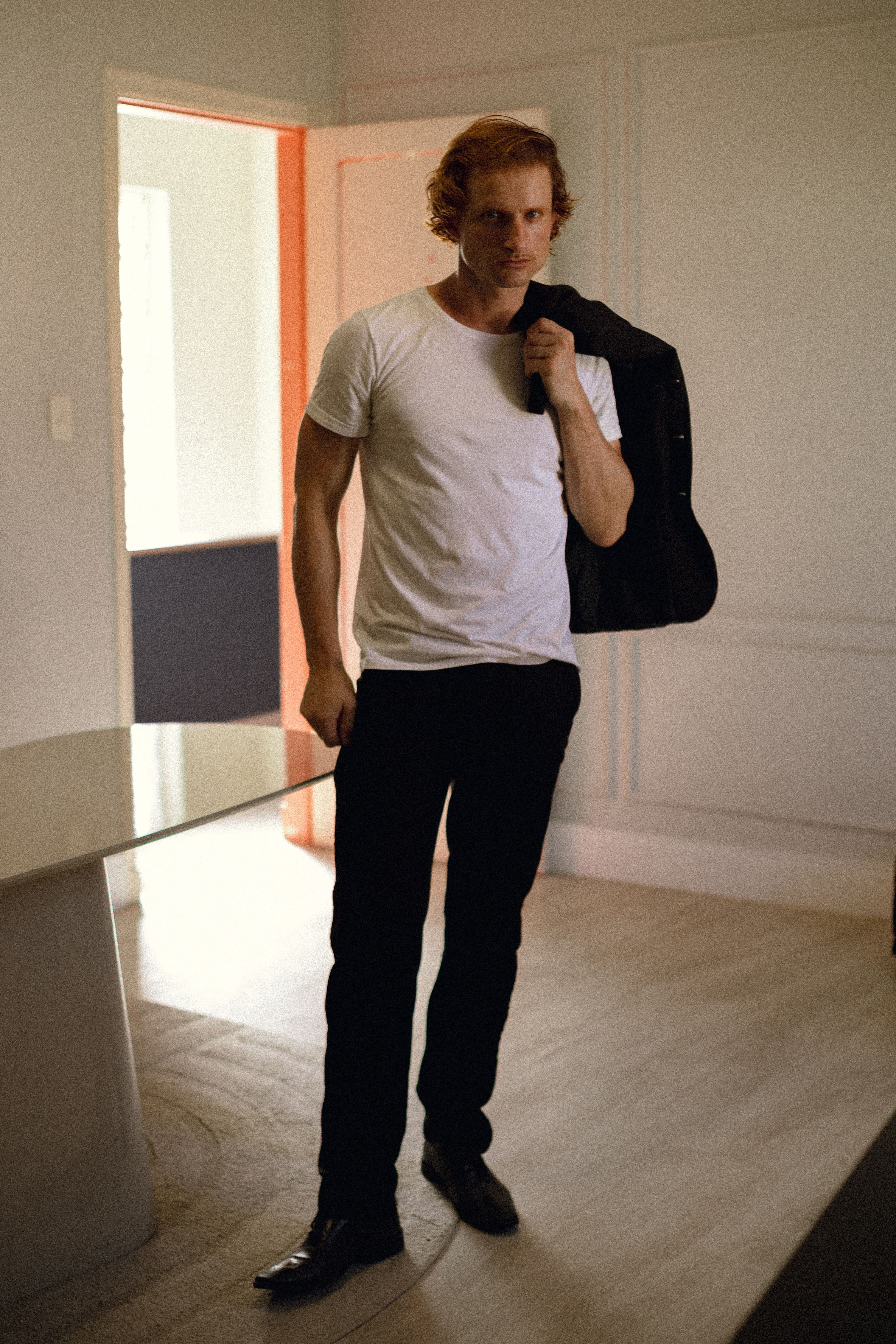 A stylish man in a white shirt and black pants in a modern interior space, holding a jacket.