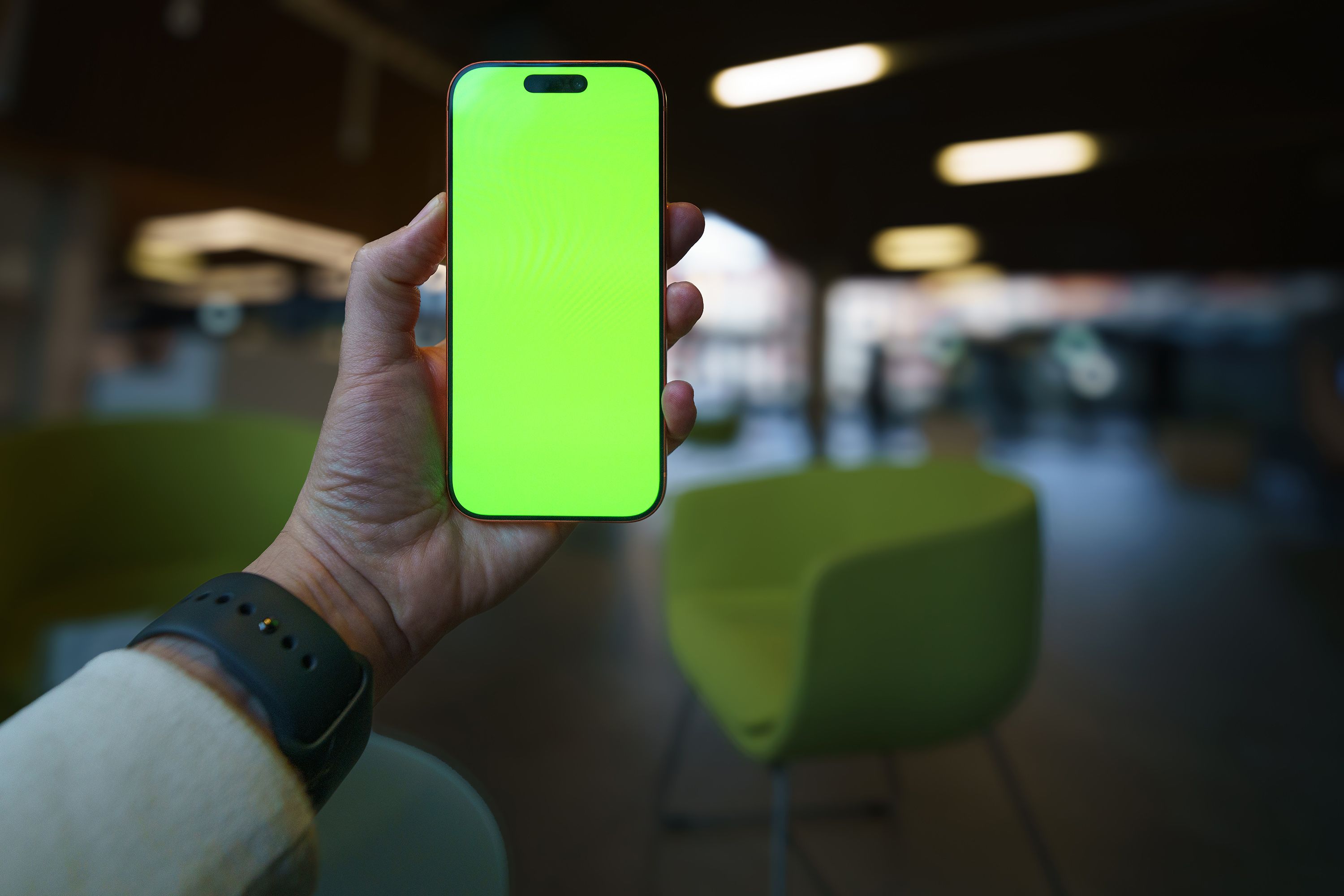 A closeup of a hand holding a smartphone with a green screen in a contemporary indoor lounge setting.