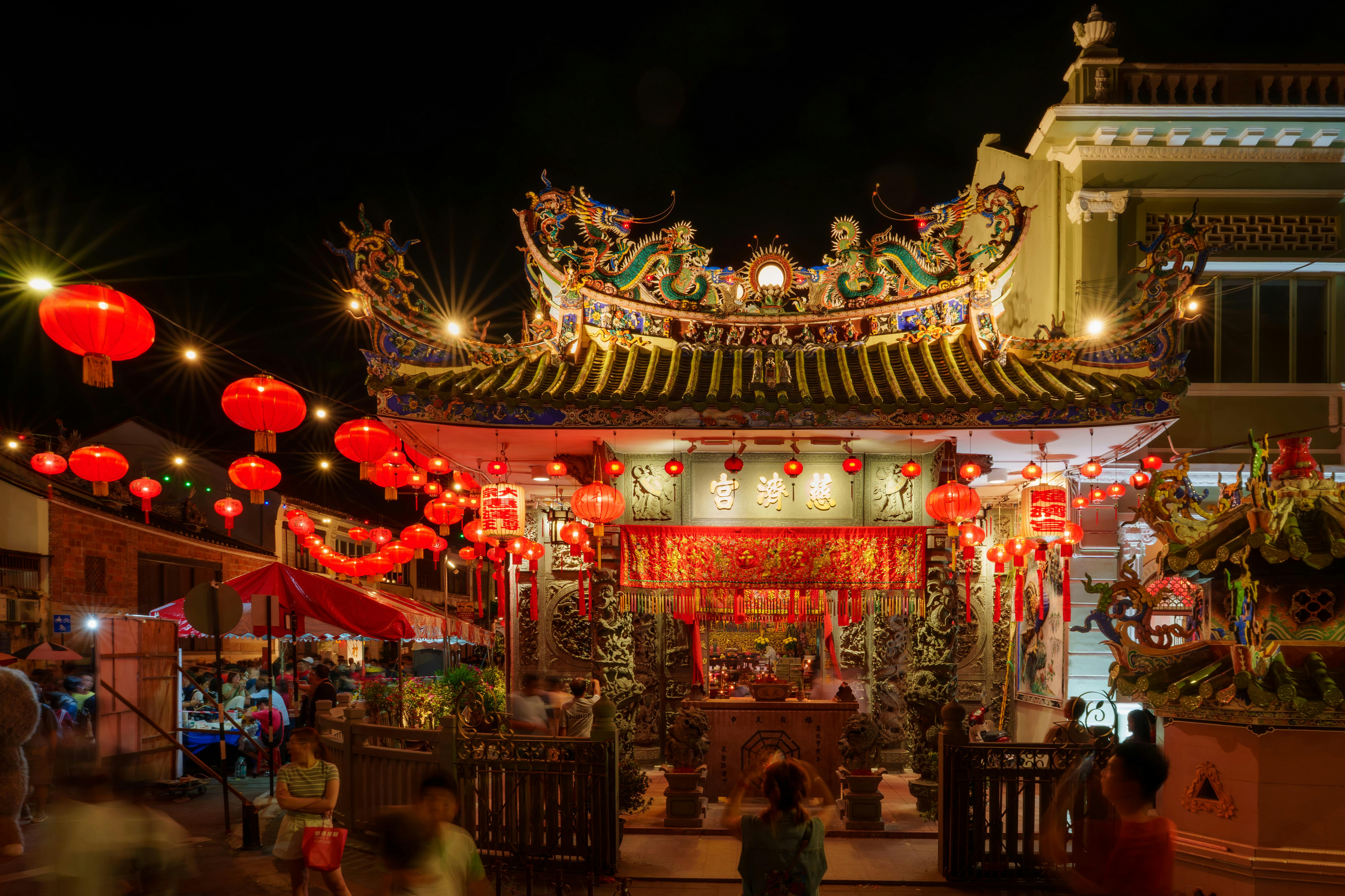 Colorful night market with red lanterns and traditional architecture, bustling crowd.