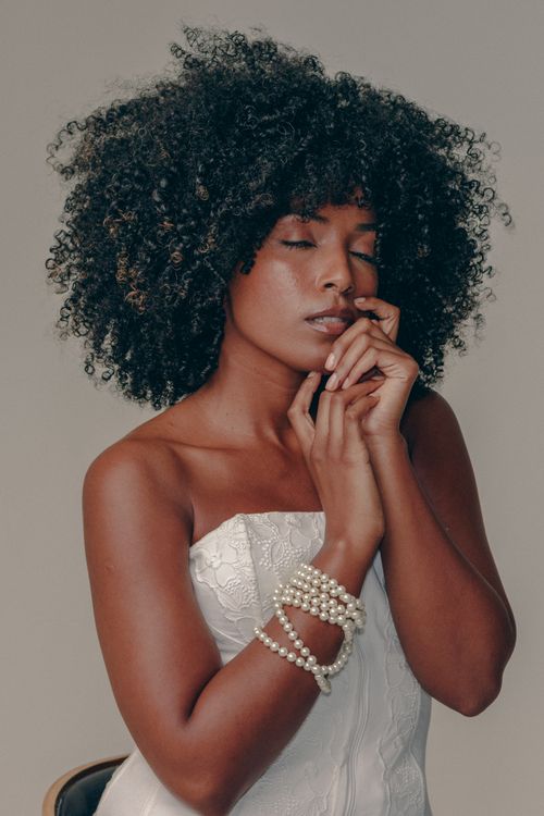 Free A woman gracefully poses in a strapless white dress adorned with pearl bracelets in a serene indoor setting. Stock Photo