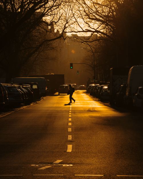Free A person crosses a city street in Berlin, silhouetted against a warm sunset glow. Stock Photo