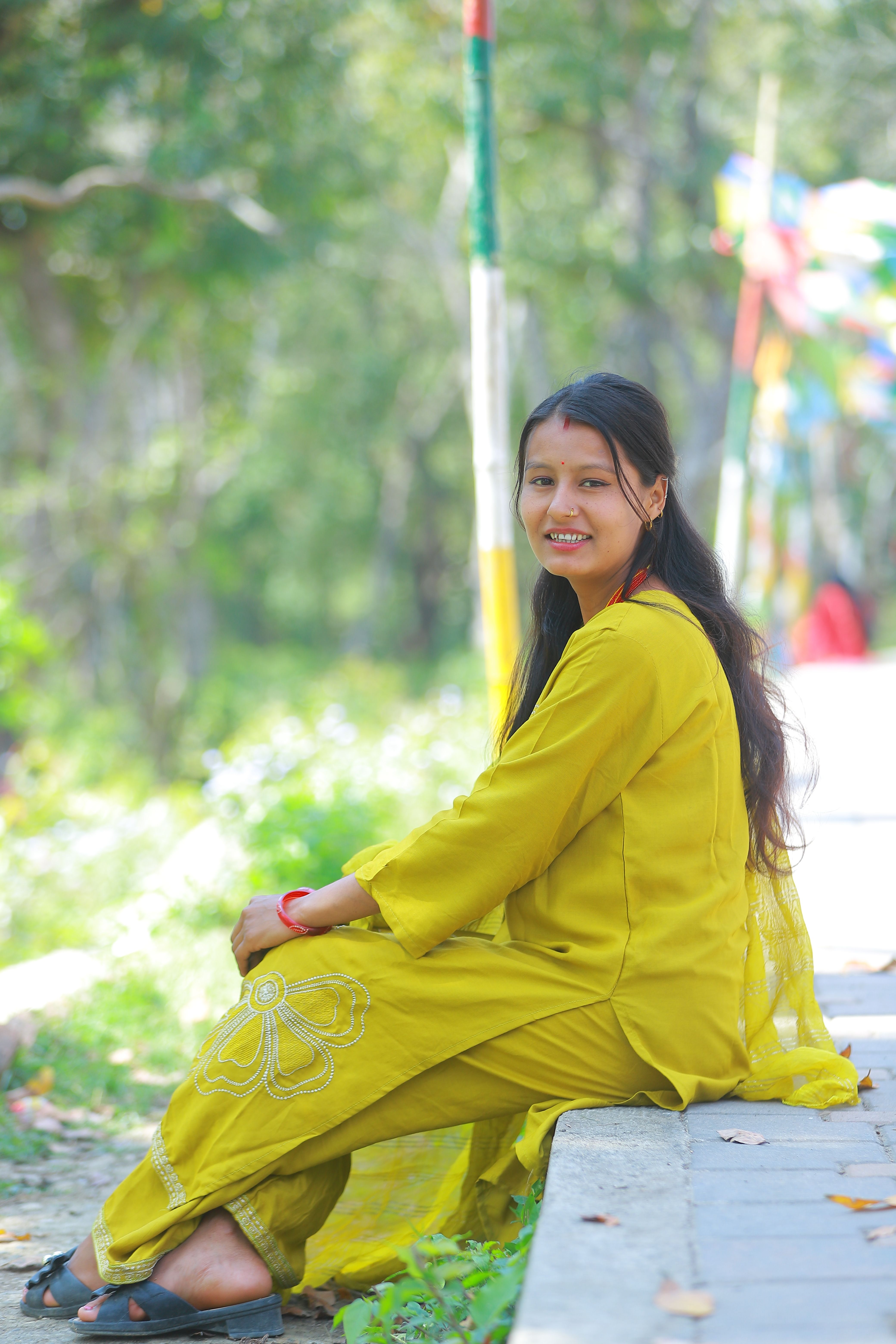 A woman in traditional yellow attire smiles while sitting in a serene outdoor setting.