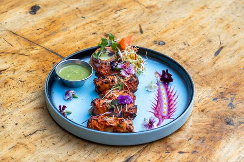 Free Gourmet tandoori chicken garnished with edible flowers and chutney, presented with artistic plating on a textured wooden table. Stock Photo