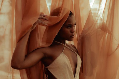 Free A stylish portrait of a woman elegantly posing behind soft orange sheer curtains. Stock Photo