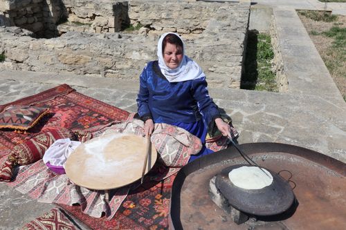 Free Woman in blue dress baking traditional bread outdoors on stone terrace with colorful carpets. Stock Photo