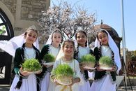 Five young girls in traditional attire holding Sabzeh, celebrating Nowruz outdoors under cherry blossoms.