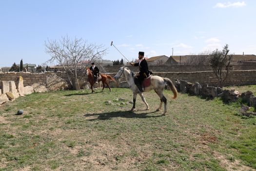 Two men on horseback engage in a traditional rural game under a clear blue sky.