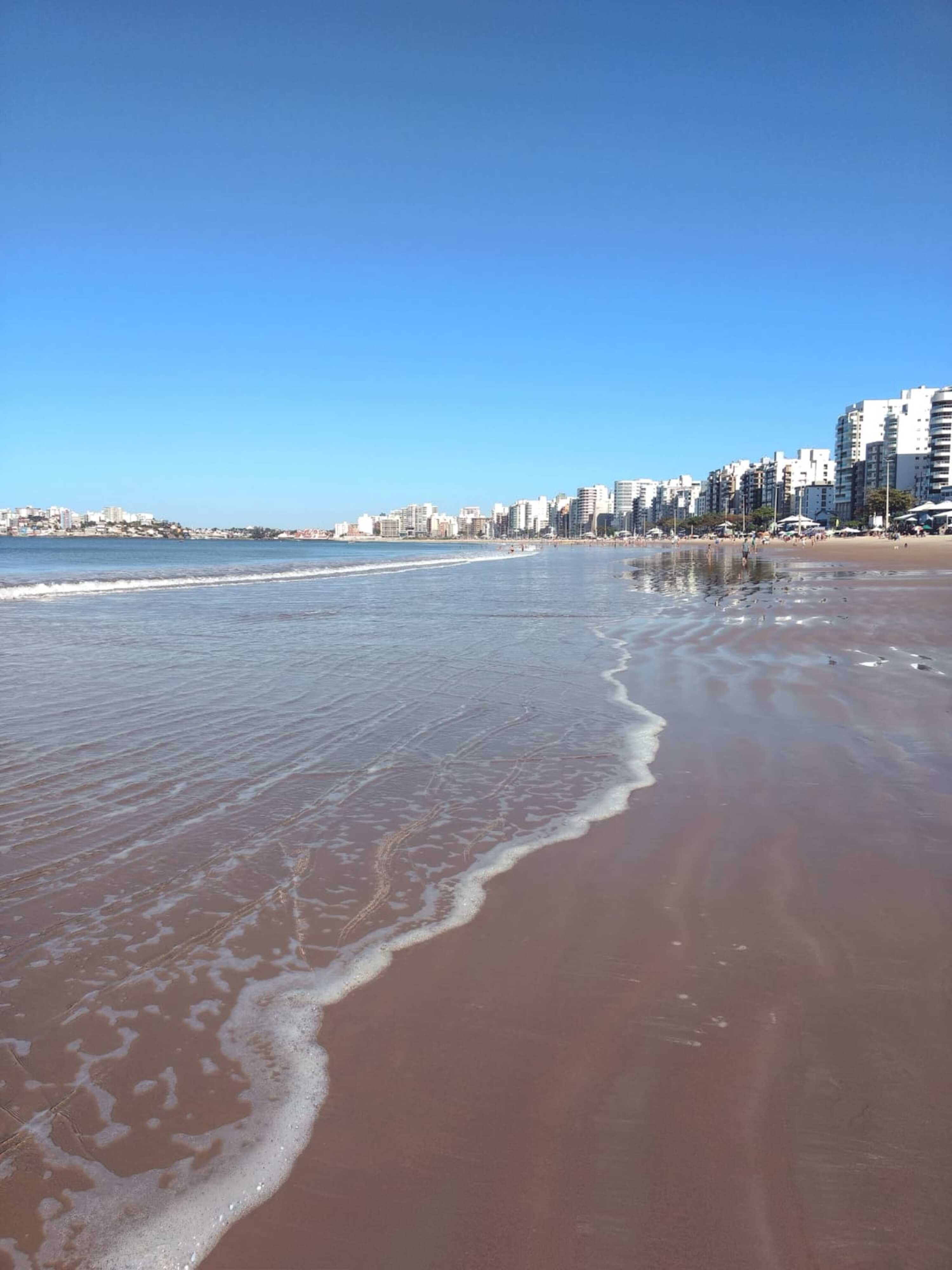 Serene view of Praia do Morro in Guarapari, featuring ocean and city skyline under a bright blue sky.
