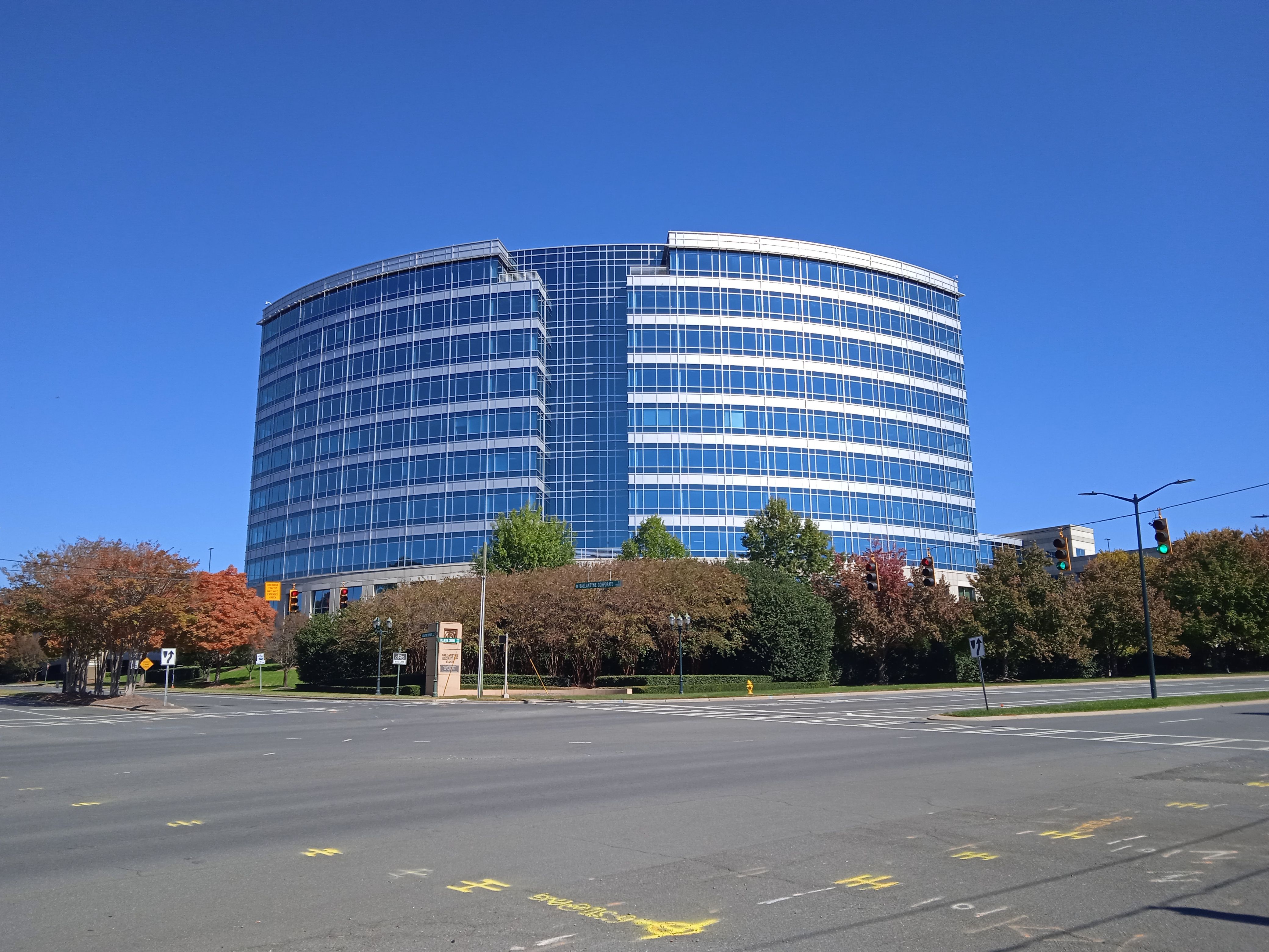 A curved glass office building with a blue sky backdrop, surrounded by autumn trees.