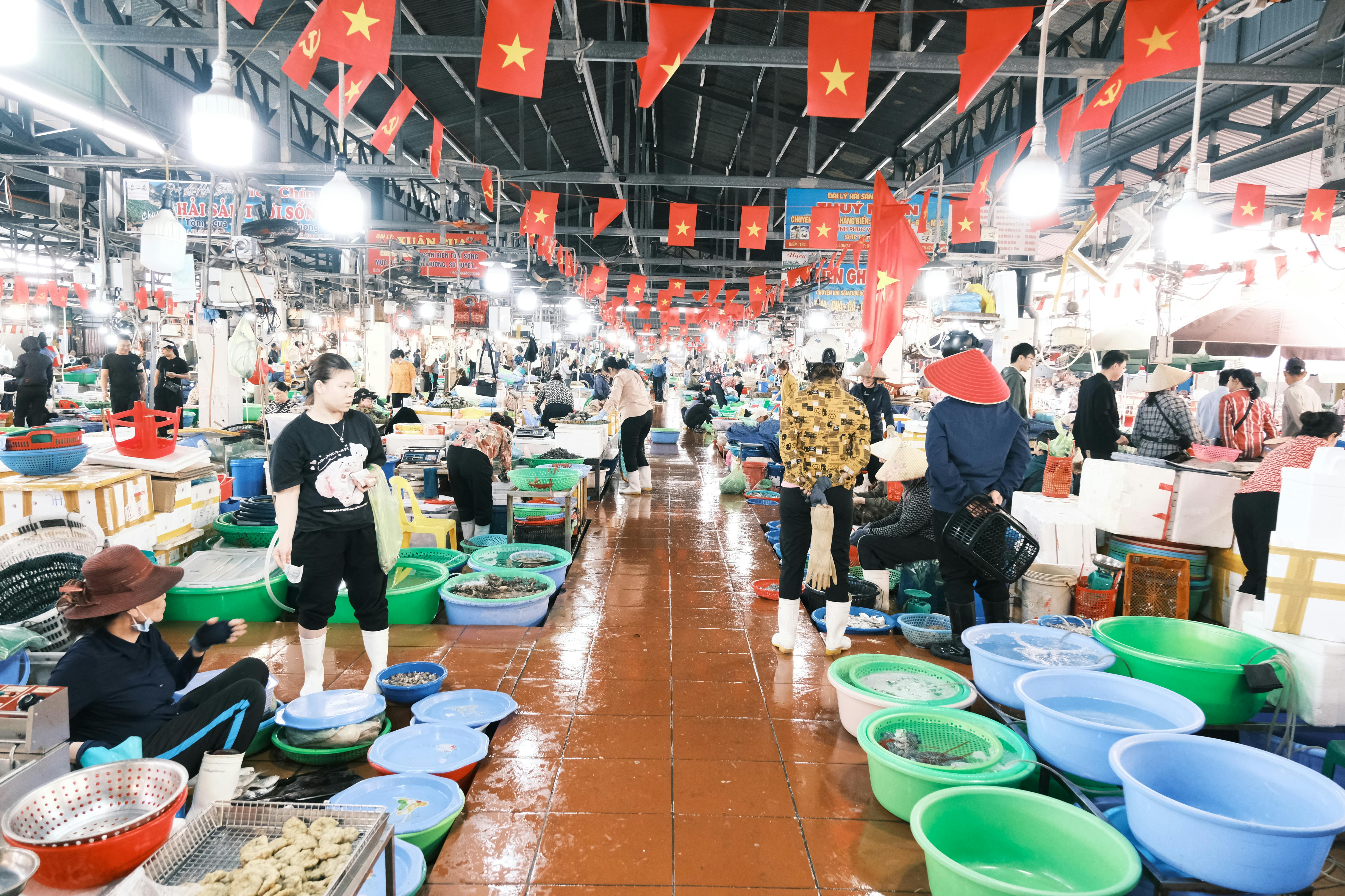 Busy indoor Vietnamese fish market bustling with customers and vendors under national flags.