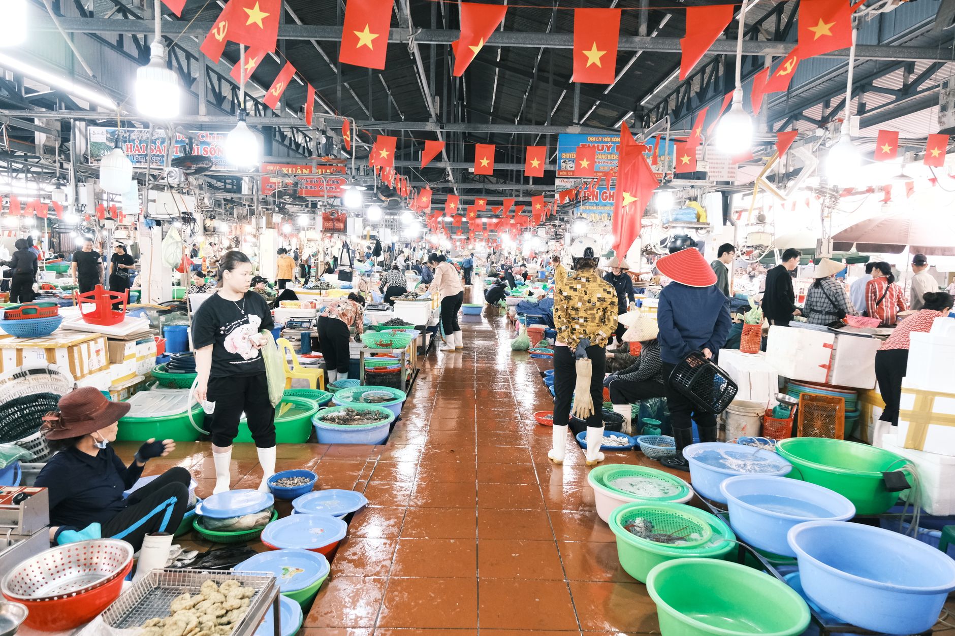 Busy indoor Vietnamese fish market bustling with customers and vendors under national flags.