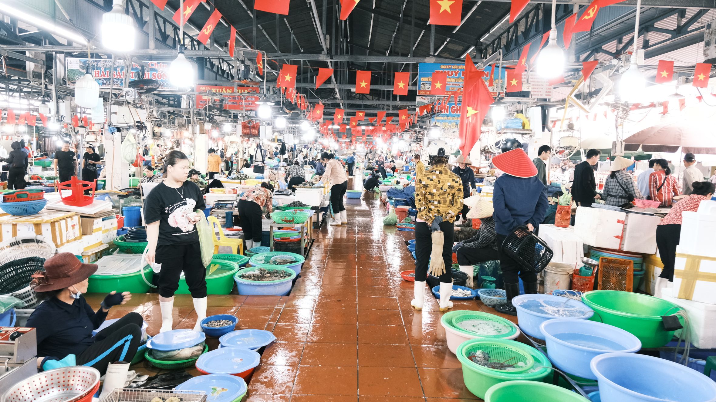 Busy indoor Vietnamese fish market bustling with customers and vendors under national flags.