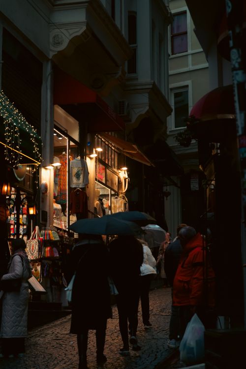 Free People with umbrellas walk through a dimly lit city alleyway lined with shops. Stock Photo
