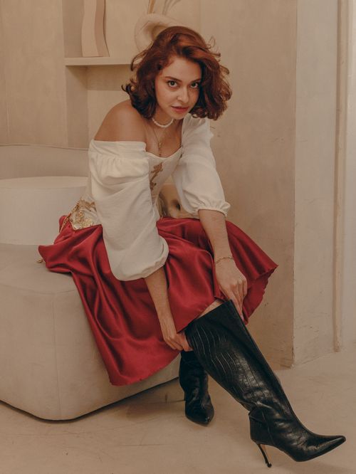 Free A fashionable woman in a red skirt and white blouse adjusts her black boots indoors. Stock Photo