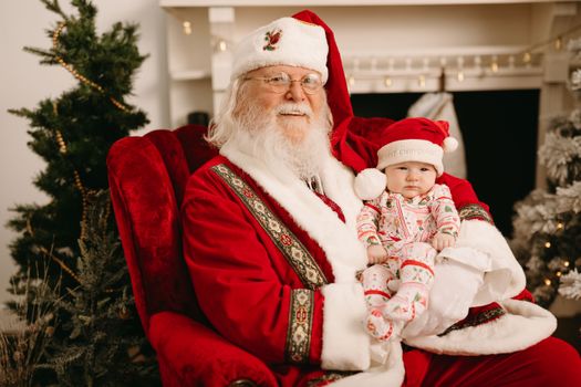 Santa Claus holding a baby dressed in festive attire, capturing the spirit of Christmas indoors.