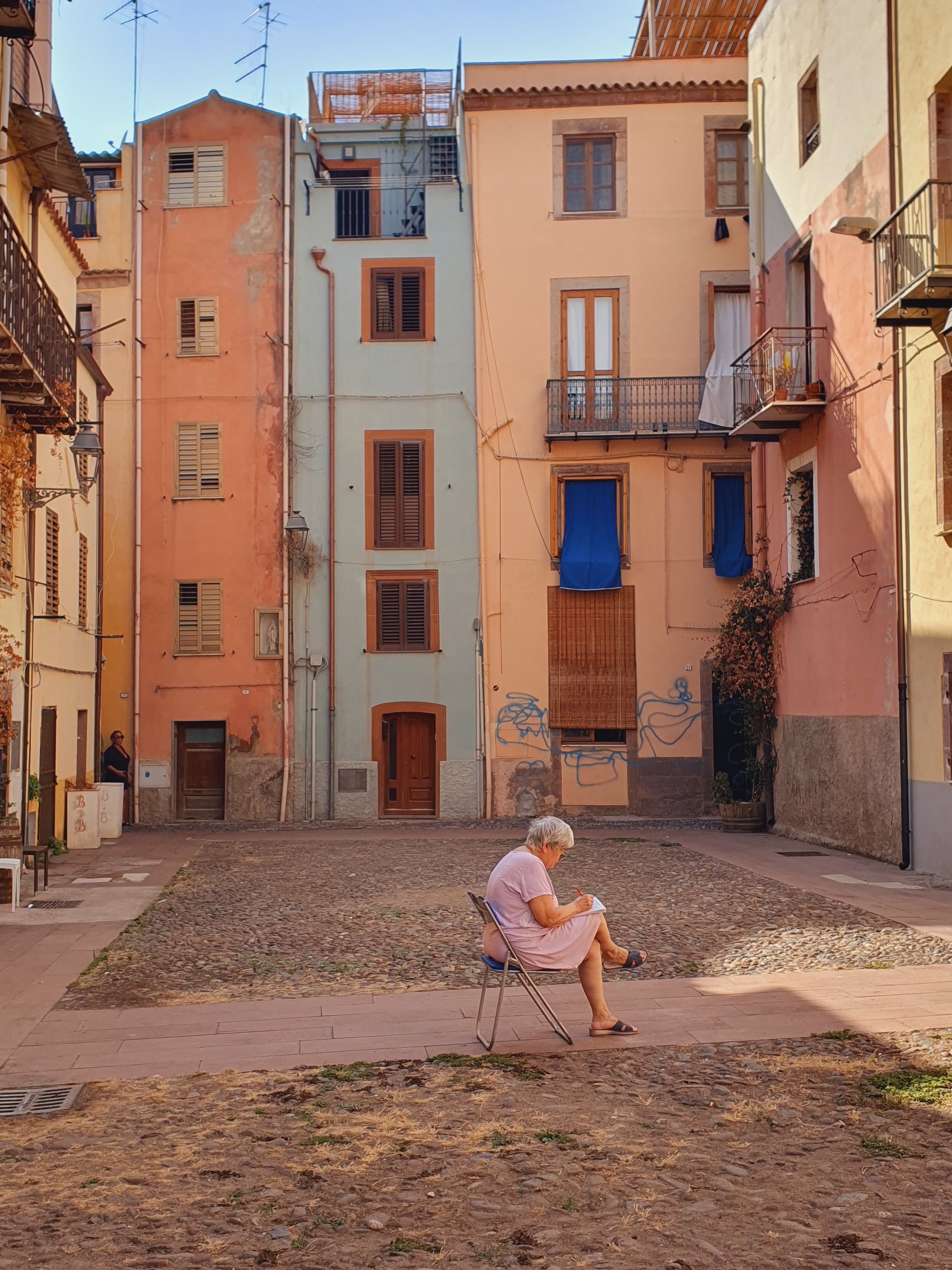 A senior woman sits reading in a picturesque European courtyard, surrounded by colorful buildings on a sunny day.