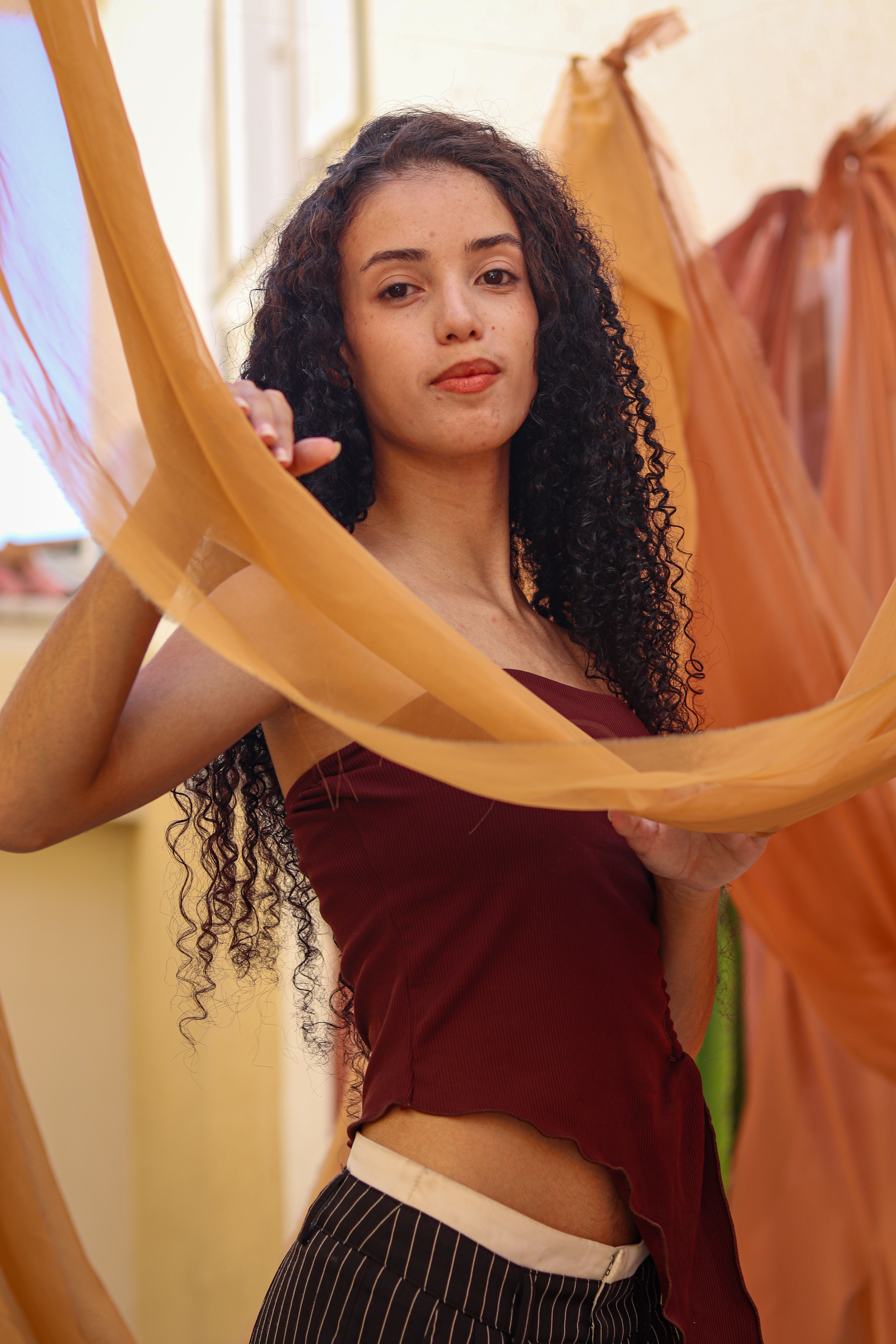 A stylish woman with curly hair poses gracefully among orange and beige curtains in a vibrant setting.