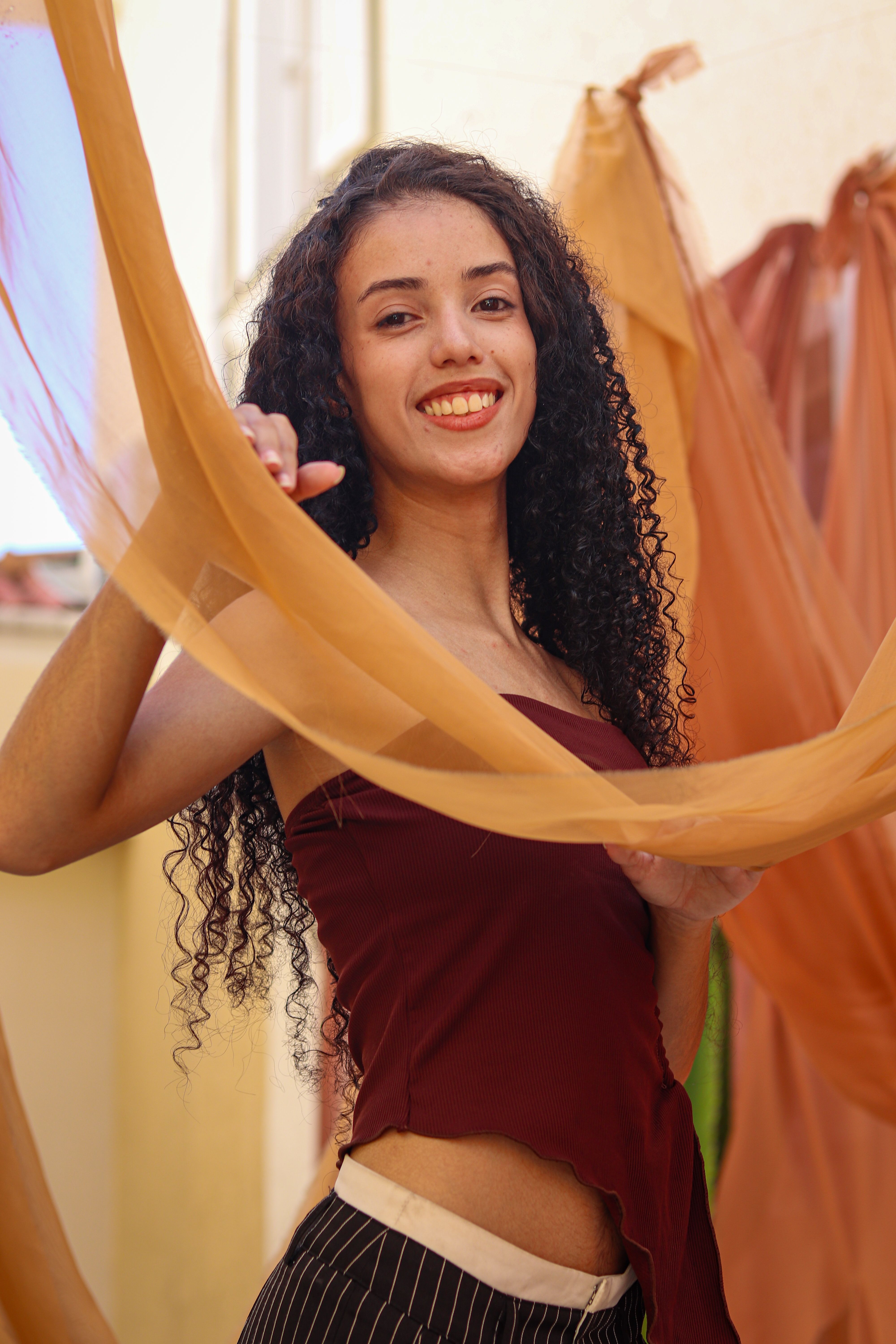 A joyful young woman with curly hair posing with colorful drapes indoors.