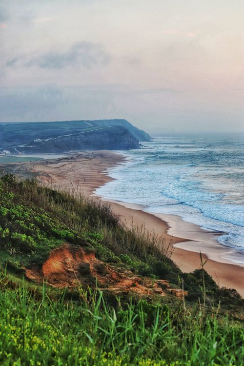 Free Stunning coastal view of waves hitting sandy shores with lush green cliffs during dawn. Stock Photo