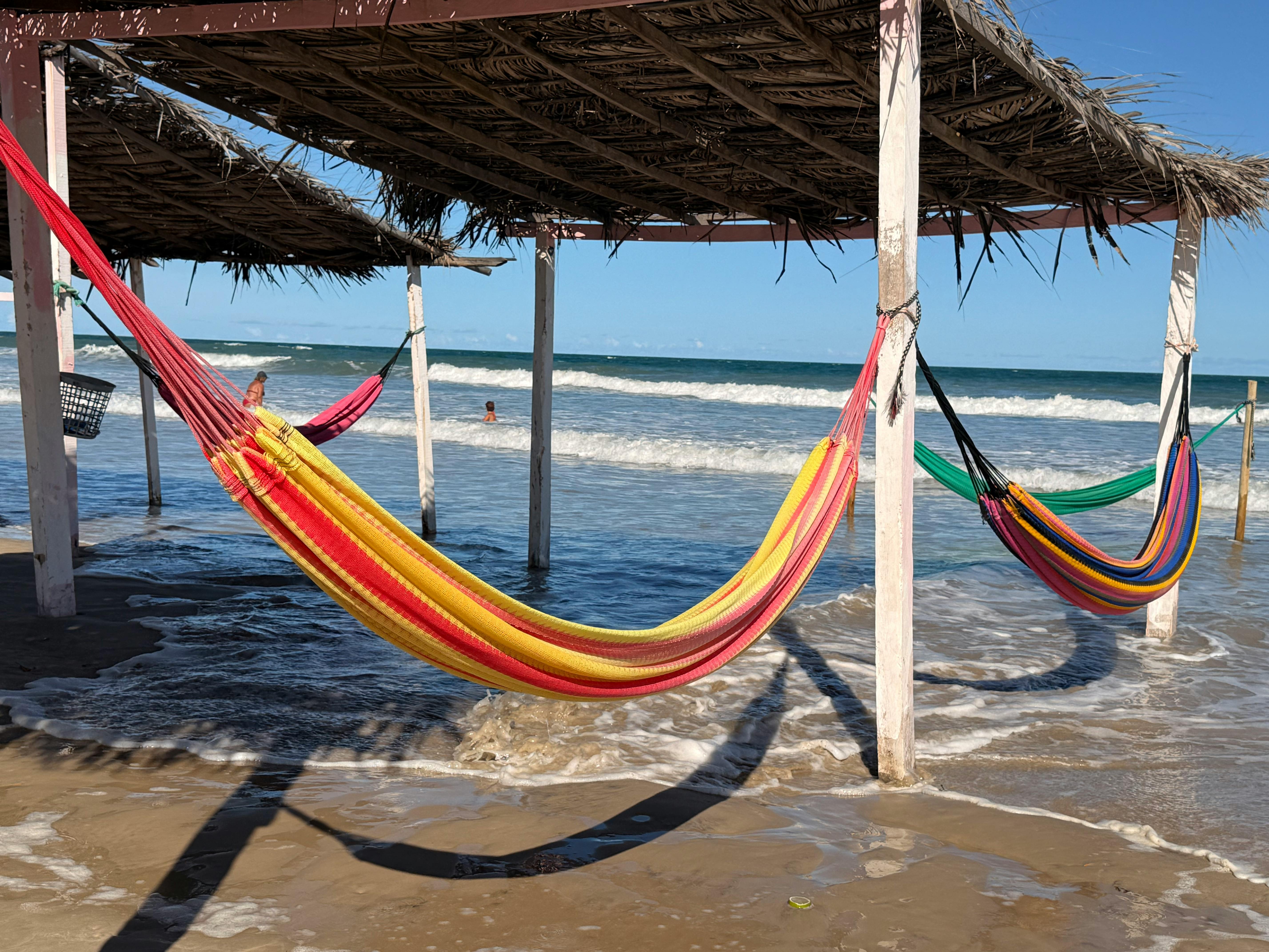 Vibrant hammocks hang under rustic shelter by ocean waves, creating a tropical vibe.