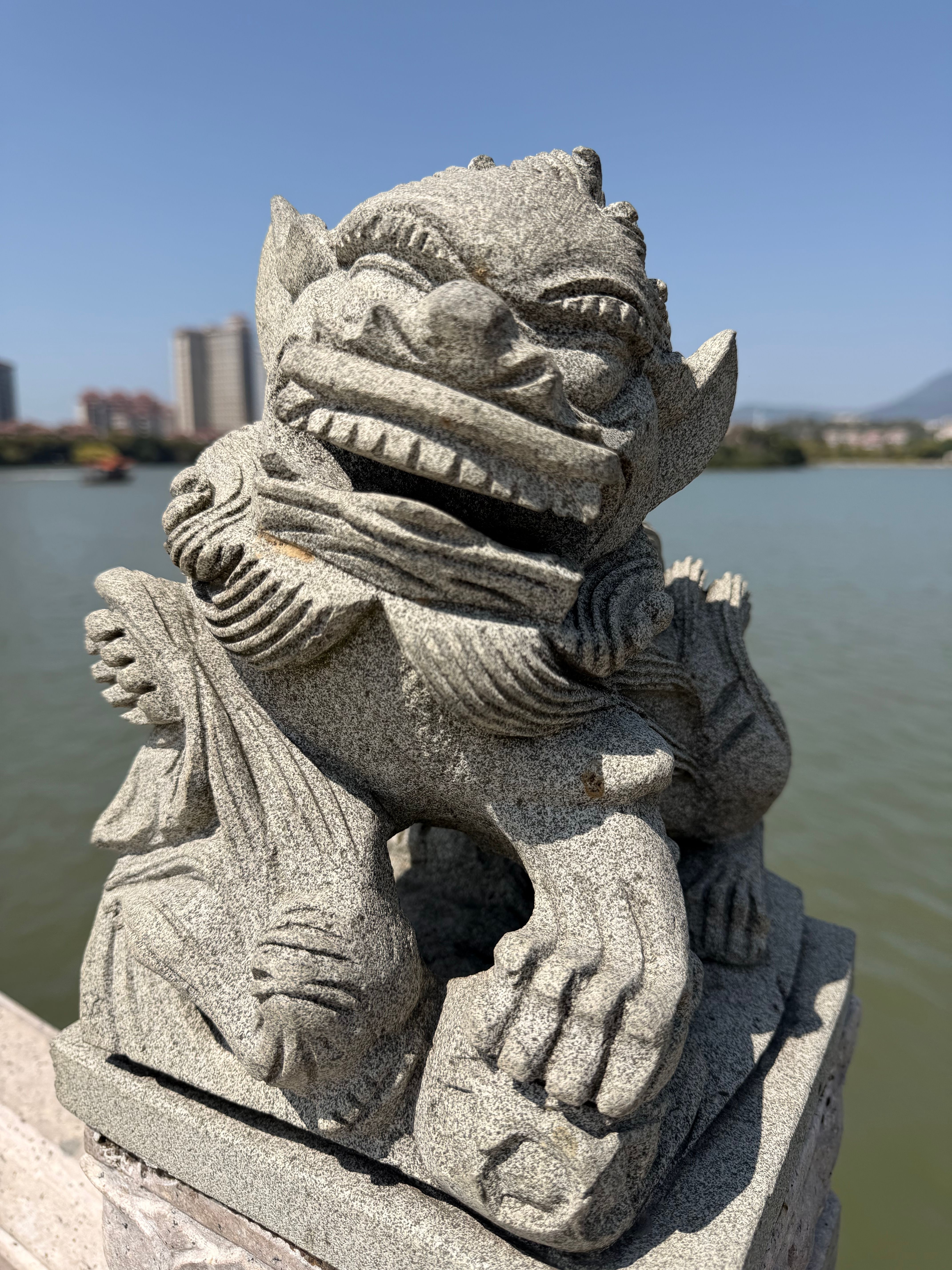 Gratuit Statue de lion gardien en pierre surplombant l'eau à Xiamen, Fujian, Chine, avec la silhouette de la ville en arrière-plan. Photos