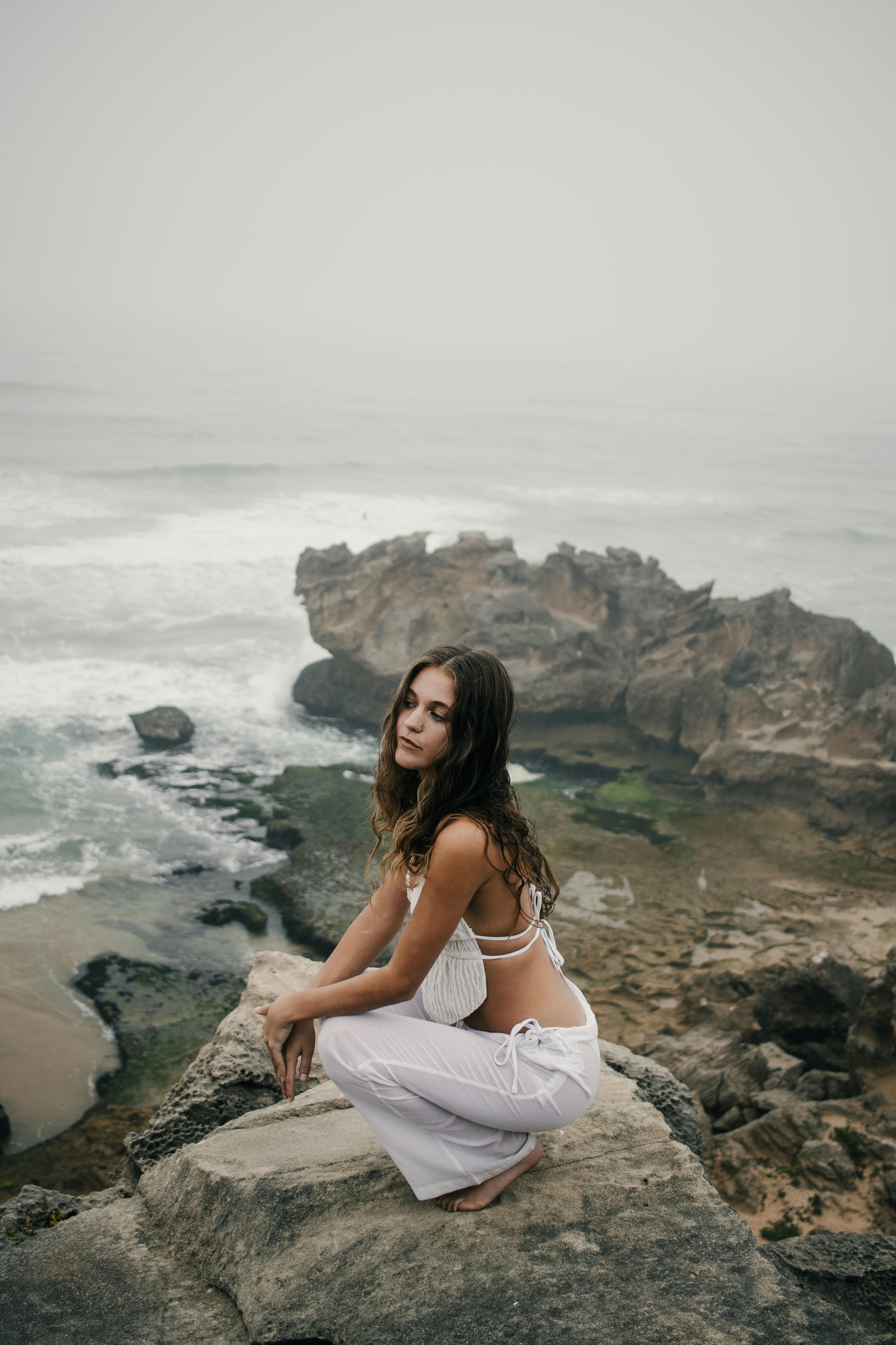A serene scene of a young woman sitting on a rocky beach with overcast skies and waves in the background.