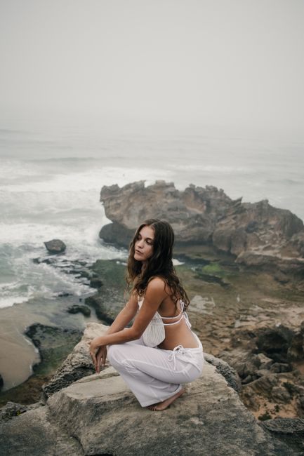 A serene scene of a young woman sitting on a rocky beach with overcast skies and waves in the background.