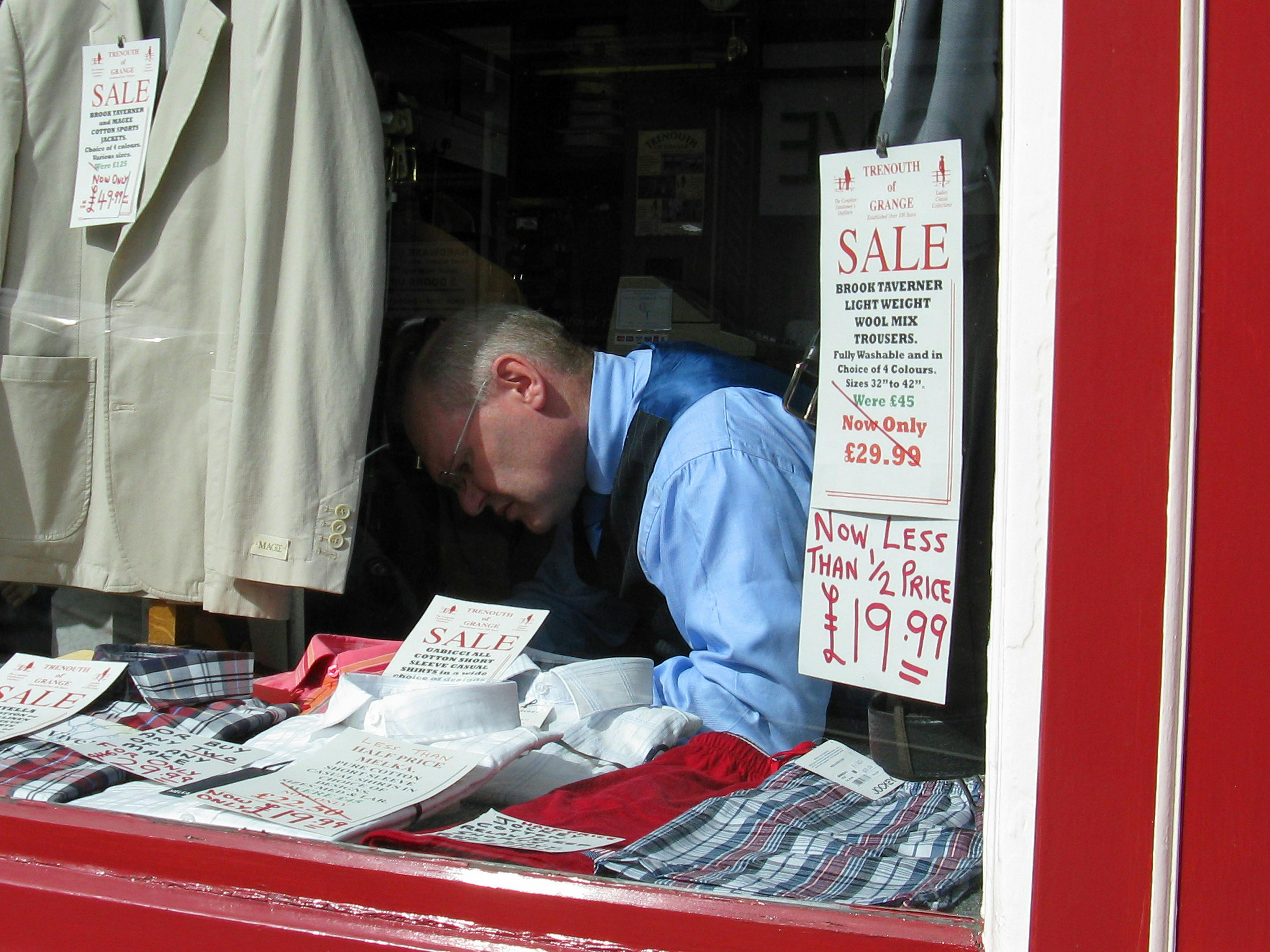 A man in a shop window arranging clothes for a sale with various sale signs displayed.
