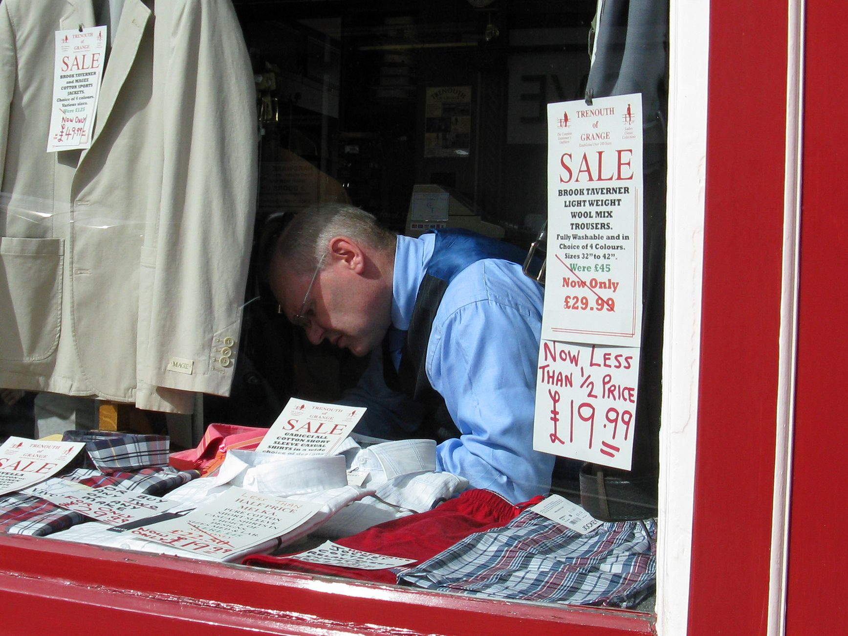 A man in a shop window arranging clothes for a sale with various sale signs displayed.