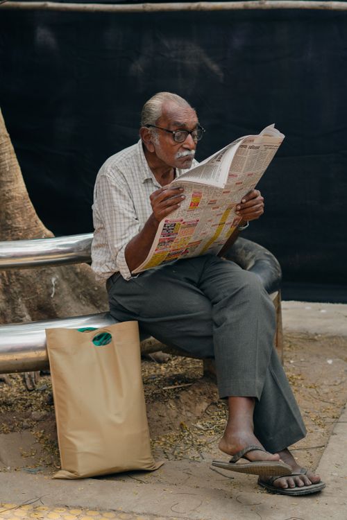 Free Senior man reading a newspaper on a bench next to a tree, enjoying a quiet moment outdoors. Stock Photo