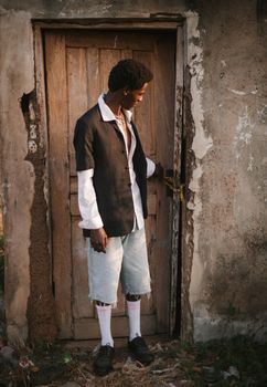 Fashionably dressed man stands at a rustic wooden door in an outdoor setting, captured in warm tones.