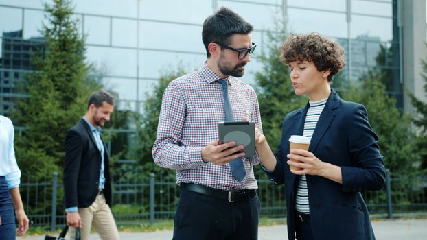 Two business professionals in conversation holding a tablet and coffee outdoors.