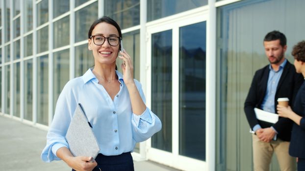 Confident businesswoman talking on phone with colleagues outside office building.