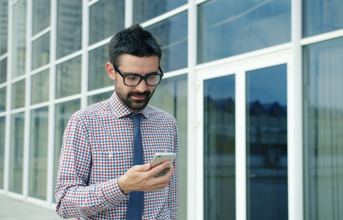 Businessman in glasses checks smartphone outside modern office building.