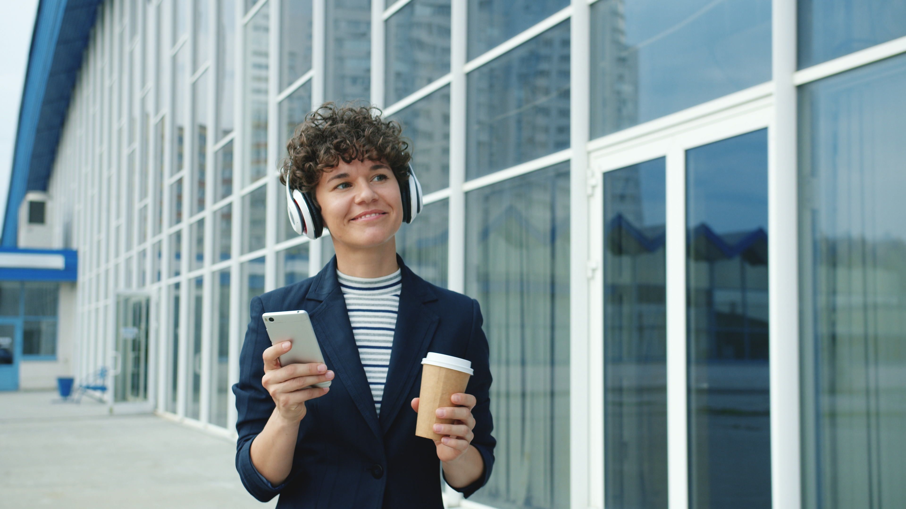 Smiling woman with headphones and coffee outside modern glass building.