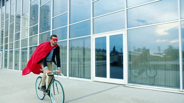 Adult man in red cape riding a bicycle past a modern glass building during the day.