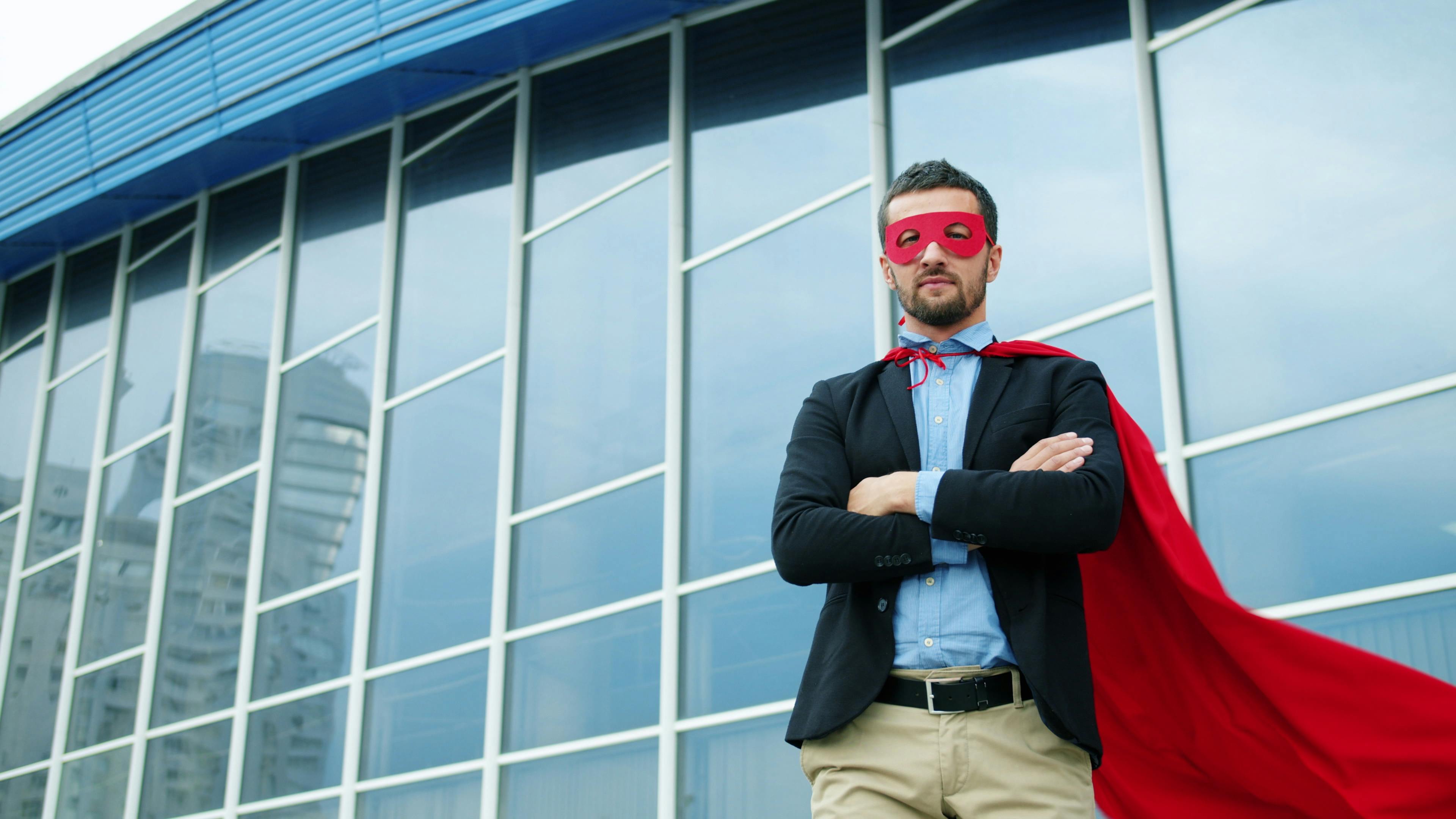 Confident businessman wearing a red superhero costume stands outside a glass building.