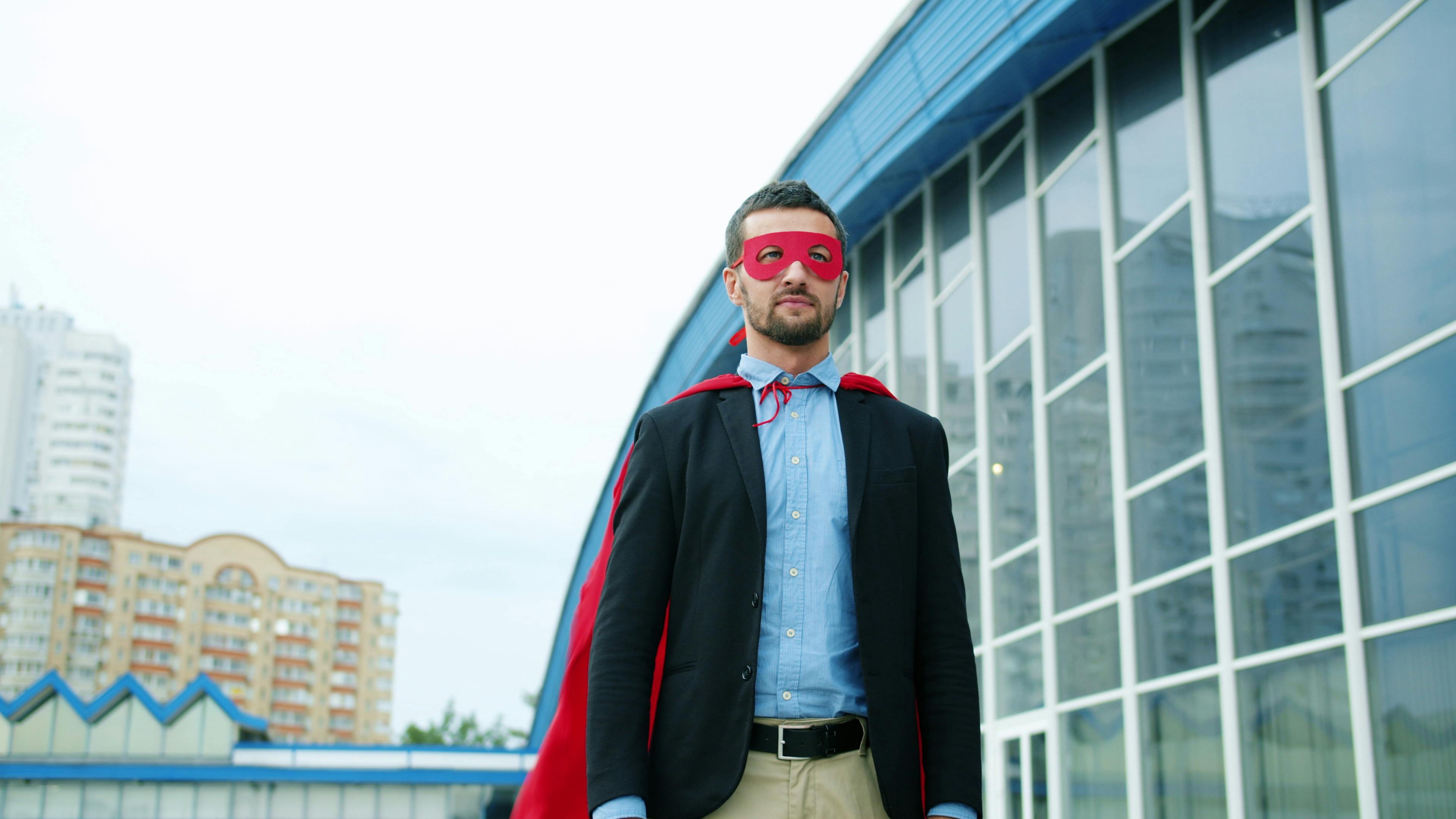 Confident businessman wearing a red superhero mask and cape standing outside a modern building.