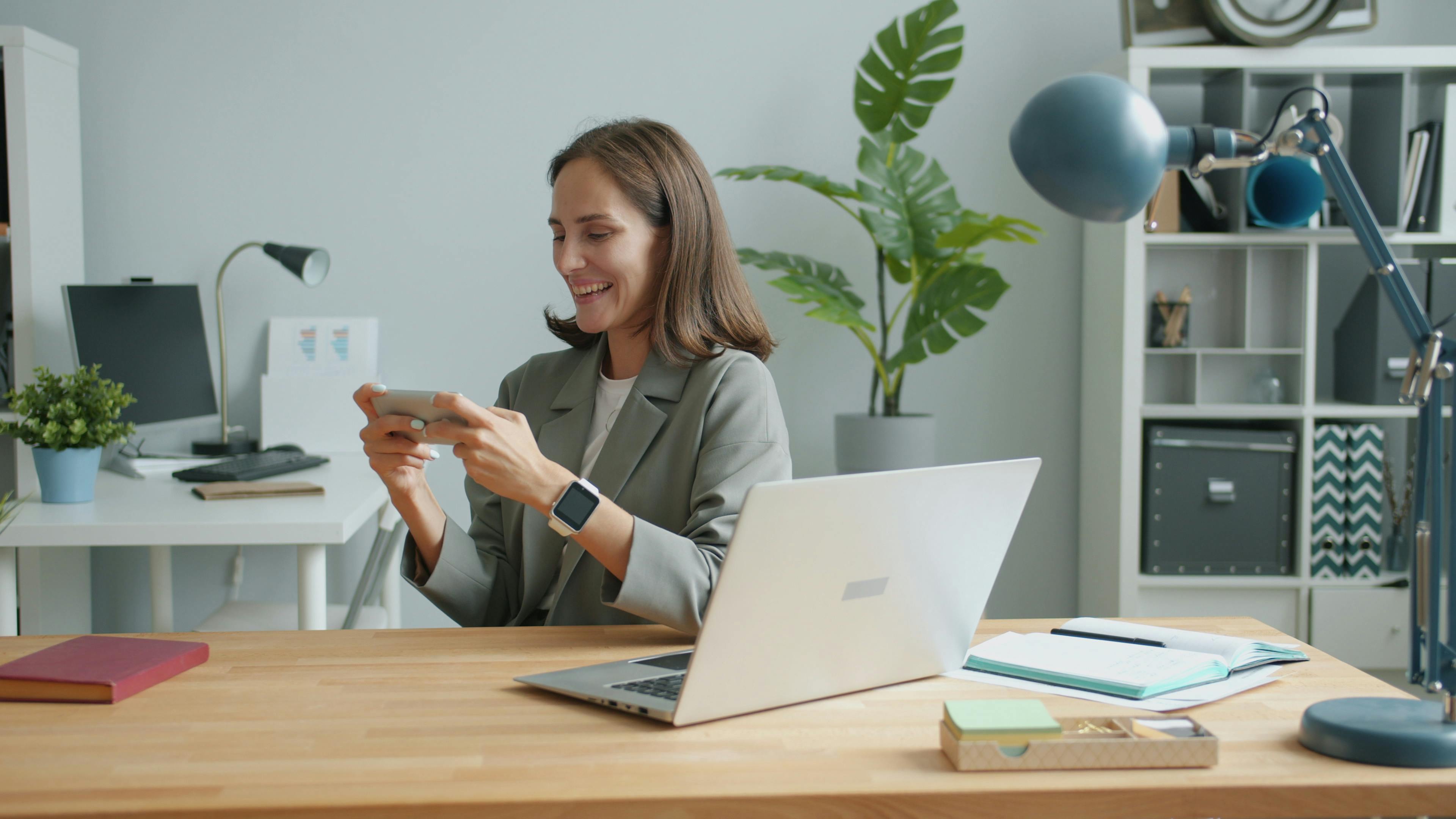 Smiling woman using a smartphone and laptop in a modern office setting.