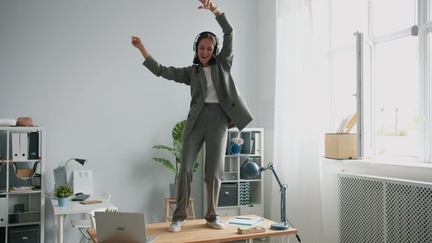 Energetic woman in suit dances on a desk in her modern office space.