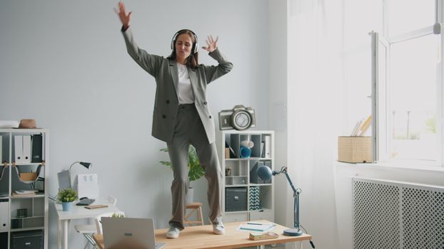 Businesswoman in suit and headphones dancing energetically on office desk.