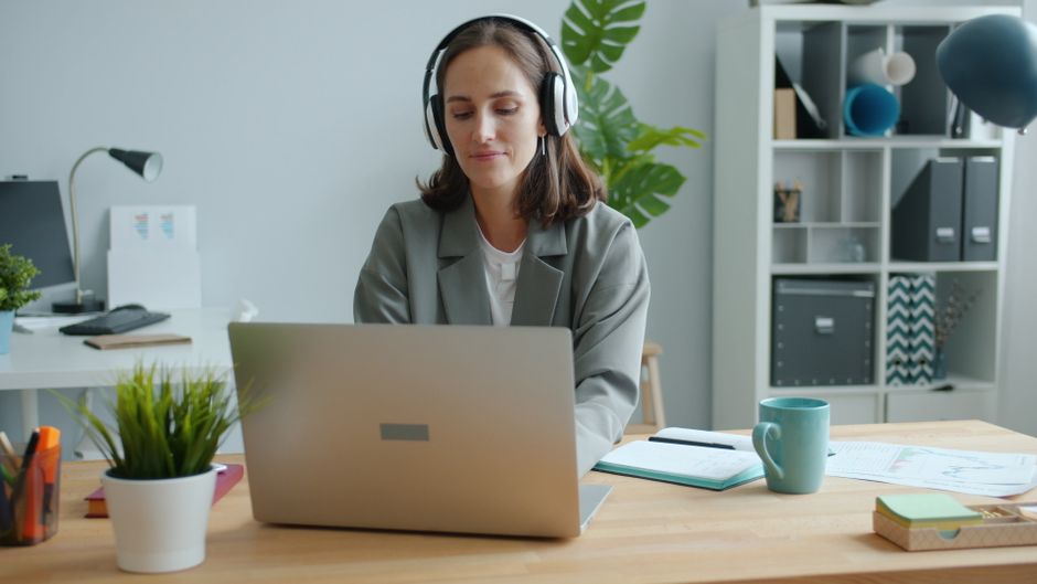 Mujer en una oficina moderna en casa usando un portátil con auriculares, trabajo concentrado.