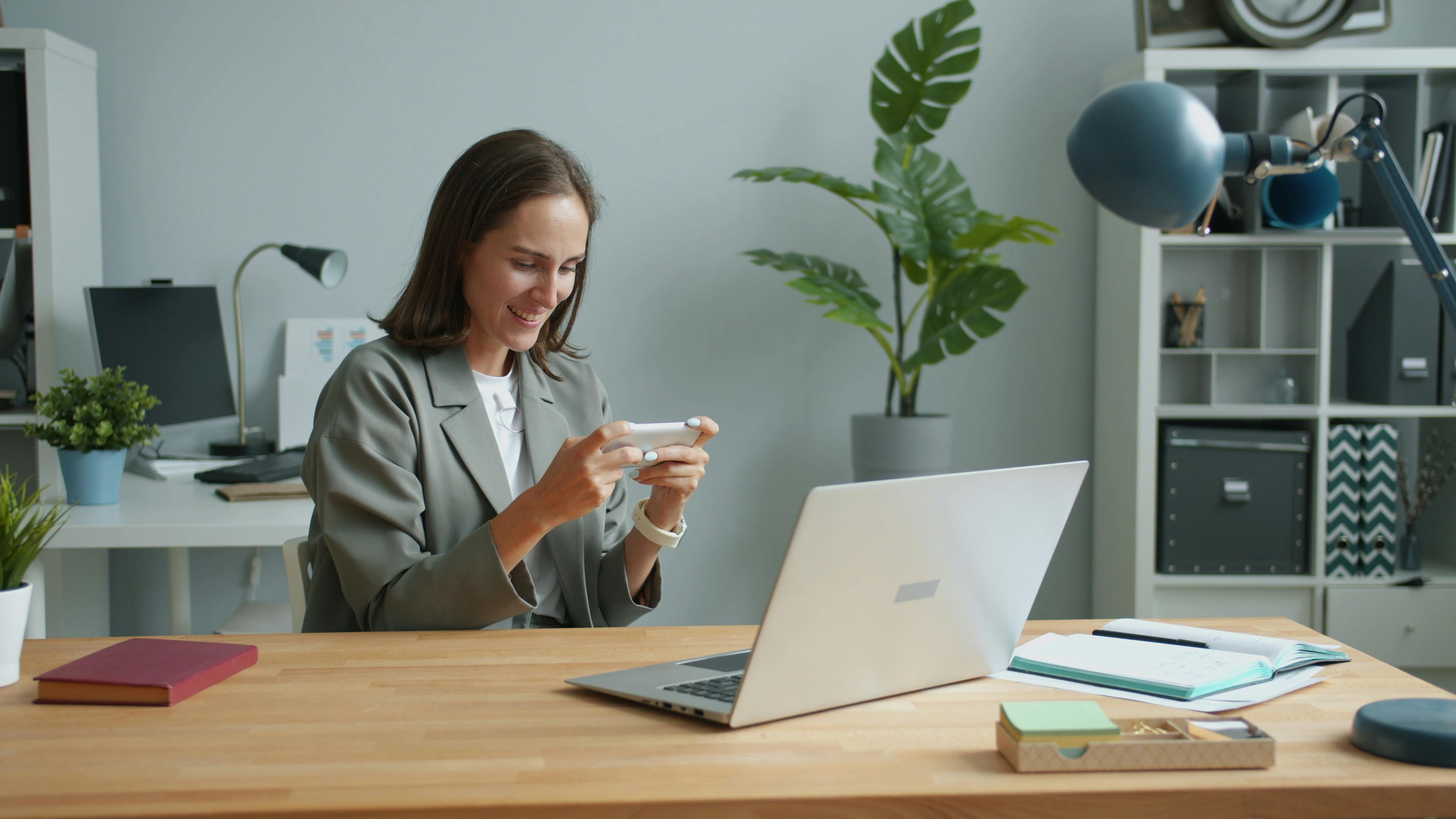A woman in a home office using a smartphone while working on a laptop.