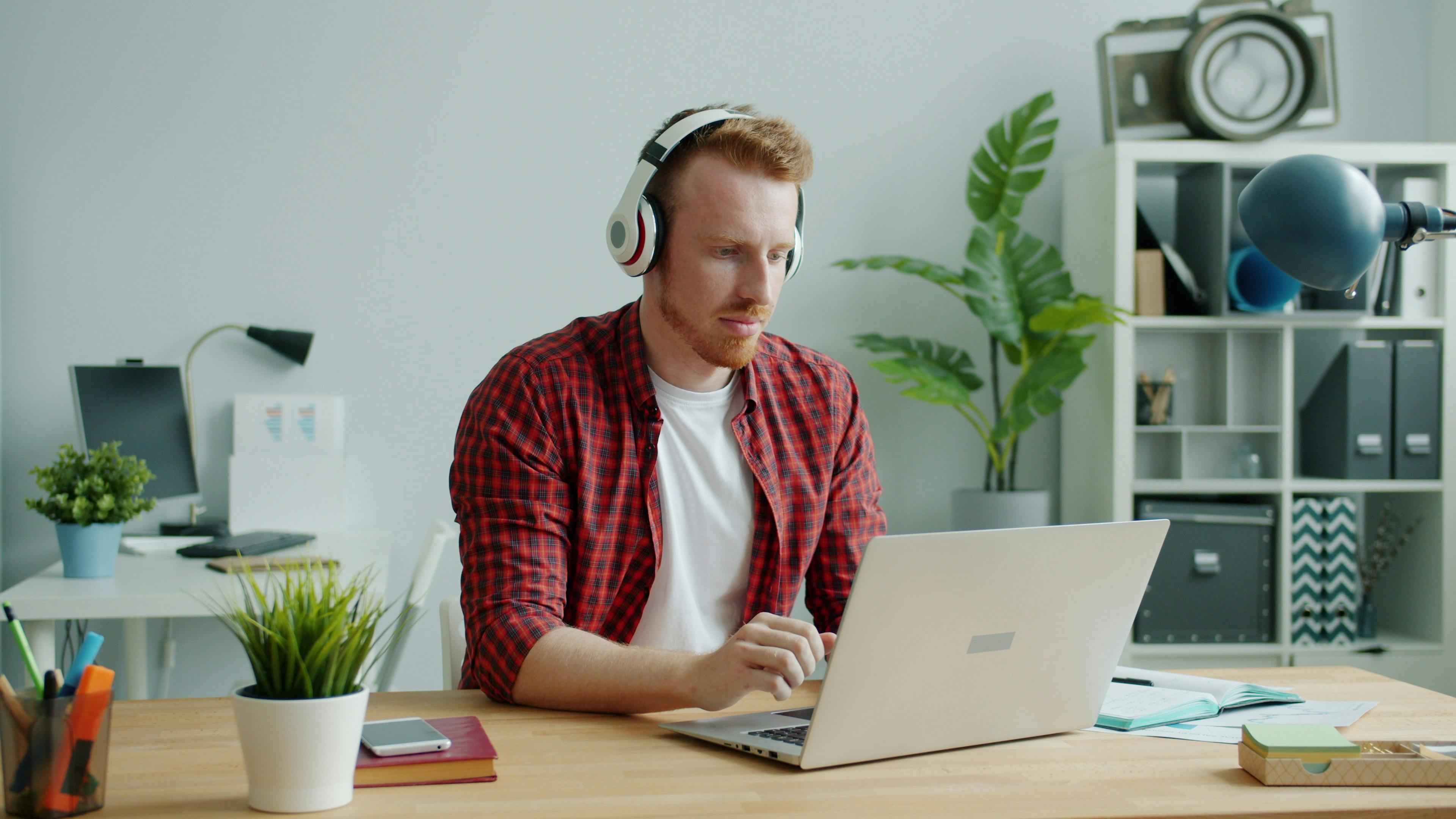 A young man using a laptop and headphones at a home office desk, surrounded by plants and decor.